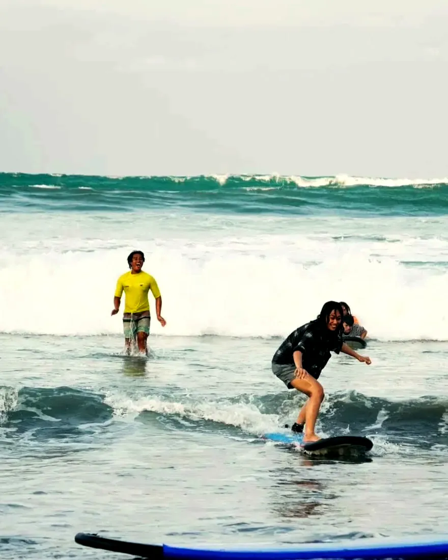 A student standing up on her board at Sabang Beach while her El Dawn instructor cheers behind her